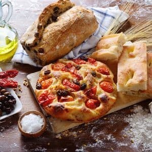 Un'ampia selezione di pani italiani, inclusi focaccia e pane alle olive, con ingredienti come olio d'oliva, pomodori e farina, su un tavolo di legno.