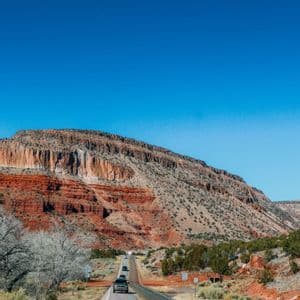 Des voitures roulent sur une route sinueuse, longeant une grande montagne de roche rouge stratifiée sous un ciel bleu éclatant.