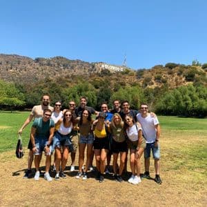 Un viaje en grupo de WeRoad posando para una foto en un parque con césped, con el letrero de Hollywood en una colina al fondo bajo un cielo despejado.