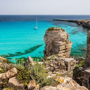 Vista da una scogliera rocciosa su una baia turchese, con una barca a vela vicino a un'imponente formazione rocciosa.