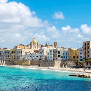 Una città costiera con edifici colorati si erge dietro un muro di pietra, affacciata su un mare turchese e una spiaggia sabbiosa sotto un cielo azzurro.