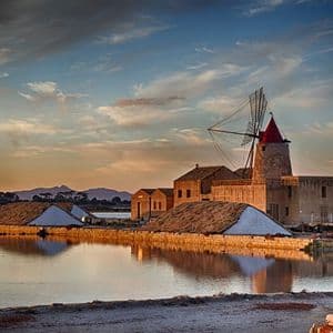 Un mulino a vento tradizionale con tetto rosso si erge accanto alle saline sul bordo dell'acqua al tramonto, con montagne in lontananza.
