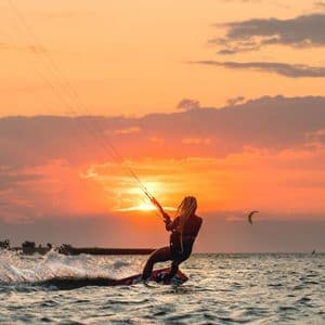 Una persona con lunghe trecce fa kitesurf sul mare, stagliata contro un vibrante tramonto arancione.
