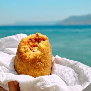 Una mano tiene un arancino di riso fritto ripieno di carne in un tovagliolo, con il mare blu sullo sfondo.
