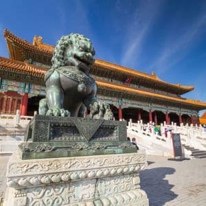 Une statue de lion gardien en bronze sur un piédestal, devant un palais chinois traditionnel, sous un ciel bleu clair.