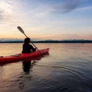 Una persona vista di spalle pagaia su un kayak rosso su acque tranquille, osservando il tramonto sulle montagne distanti.