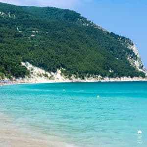 Una spiaggia sabbiosa affollata ai piedi di una montagna alberata, con acqua turchese che lambisce la riva sotto un cielo azzurro e limpido.