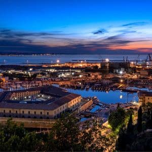 Una vista dall'alto di una grande città portuale illuminata al tramonto, con una fortezza storica nel porto.