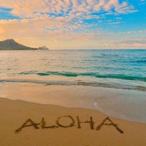 ALOHA im Sand am Strand, mit Wellen und Bergblick bei Sonnenuntergang.