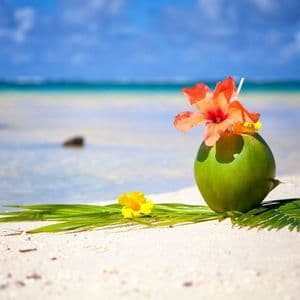 Ein Kokosnuss-Cocktail, garniert mit einer orangen Hibiskusblüte, steht an einem weißen Sandstrand mit dem Ozean im Hintergrund.