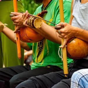Primo piano di tre persone sedute con strumenti musicali tradizionali berimbau e uno shaker caxixi.