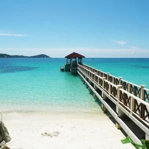 Un pontile di legno con un gazebo si estende da una spiaggia di sabbia bianca su acque turchesi limpide verso isole verdi e lontane.