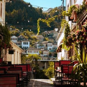 Un vicolo stretto acciottolato fiancheggiato da sedie e tavolini rossi da caffè si affaccia su un villaggio costruito su una rigogliosa collina verde sotto un cielo soleggiato.
