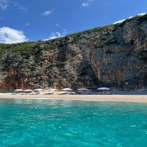 Una spiaggia sabbiosa con ombrelloni bianchi e lettini si trova alla base di un'ampia scogliera rocciosa, vista dall'acqua turchese.