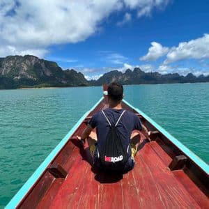 Una persona con una mochila WeRoad se sienta en la proa de un barco de madera en un lago turquesa, con montañas verdes en la distancia.