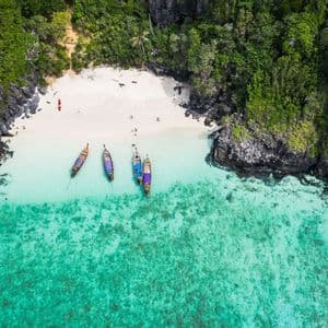 Une vue aérienne de plusieurs bateaux traditionnels amarrés dans des eaux turquoise claires le long d'une plage de sable blanc isolée, entourée de falaises verdoyantes.