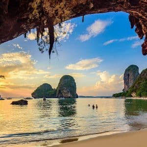 Vue depuis l'intérieur d'une grotte marine surplombant une plage tropicale avec des karsts calcaires et des bateaux au coucher du soleil.