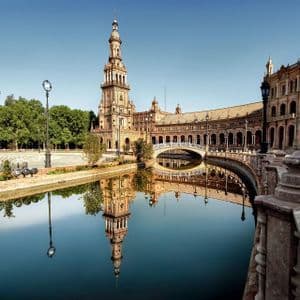 Un edificio decorato con un'alta torre si specchia in un canale tranquillo, con un ponte che lo attraversa sotto un cielo sereno.