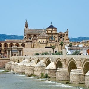 Un ponte di pietra a più arcate attraversa un ampio fiume, conducendo a una città storica con una grande cattedrale e un campanile.