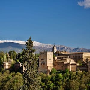 Un complesso storico di palazzi sorge su una verde collina, con una catena montuosa innevata sullo sfondo e un cielo azzurro e limpido.