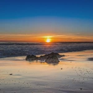 Il sole tramonta sull'oceano, la sua luce si riflette sulla sabbia bagnata di una spiaggia con scogli nell'acqua bassa.