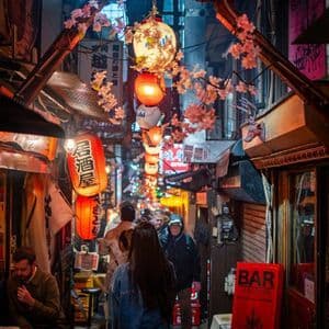 Una folla di persone cammina lungo un vicolo stretto di notte, illuminata da sfavillanti lanterne di carta rosse e bianche e fiori di ciliegio.