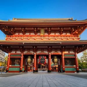 Un grande portale tradizionale giapponese rosso con una lanterna centrale appesa si erge in un cortile pavimentato sotto un cielo azzurro e limpido.
