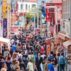 Una grande folla di persone che percorre una strada stretta, fiancheggiata da edifici con colorate insegne giapponesi.