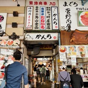 Gente che cammina e mangia in un vicolo affollato di un mercato alimentare giapponese, costeggiato da insegne di ristoranti e display di menu.