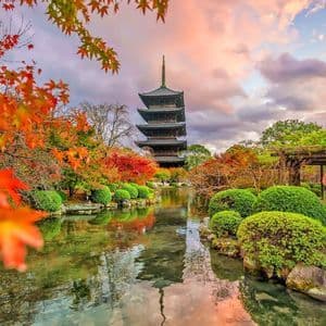 Una pagoda giapponese si specchia in uno stagno, con una persona in kimono tra il vibrante fogliame autunnale.