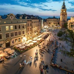 Una vista dall'alto di una vivace piazza storica al tramonto, piena di gente che passeggia e cena ai caffè all'aperto sotto gli ombrelloni.
