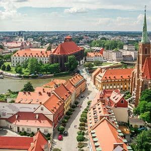 Una vista aerea di una storica città europea con tetti in tegole rosse, una chiesa imponente e un fiume che la attraversa in una giornata di sole.
