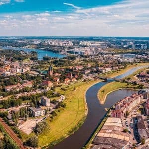 Una vista aerea di una città tentacolare dove un fiume scorre tra rigogliose aree verdi e gruppi di edifici sotto un cielo blu.