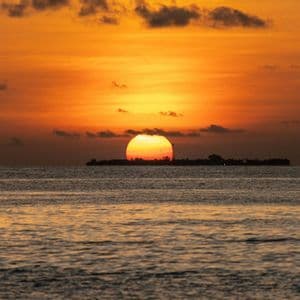 Un grande sole arancione tramonta sull'oceano, parzialmente nascosto da un'isola in silhouette sotto un cielo nuvoloso.