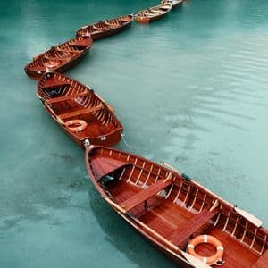 Eine Reihe aneinandergebundener leerer Holzruderboote treibt in einer geschwungenen Linie auf ruhigem, klarem türkisfarbenem Wasser.