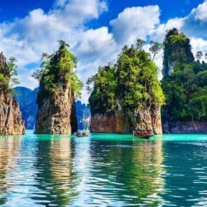 Dos barcos de cola larga flotan en aguas turquesas entre altos acantilados de piedra caliza cubiertos de árboles bajo un cielo azul con nubes blancas.