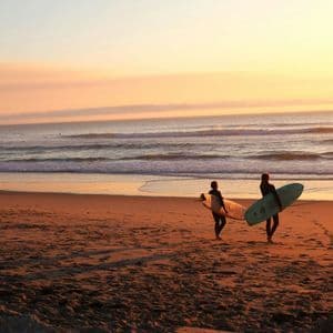 Zwei Surfer in Neoprenanzügen tragen ihre Surfbretter, während sie bei einem warmen Sonnenuntergang an einem Sandstrand entlang zum Meer gehen.