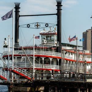 Un classico battello a vapore a pale, bianco e rosso, è ormeggiato sul fiume, con un moderno grattacielo e un grande ponte sullo sfondo.