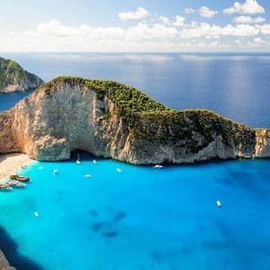 Luftaufnahme einer abgelegenen Bucht mit einem Schiffswrack an einem weißen Sandstrand, umgeben von Klippen und türkisfarbenem Wasser mit Booten.