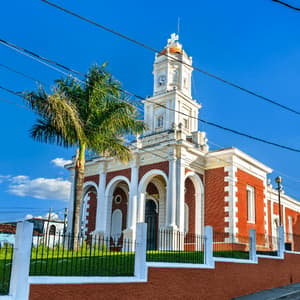 Una chiesa di mattoni rossi con un campanile bianco e dettagli ad arco si erge accanto a una palma sotto un cielo limpido e azzurro.