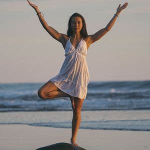 Una donna in abito bianco pratica la posizione dell'albero yoga in equilibrio su una roccia su una spiaggia al tramonto.