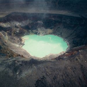 Veduta aerea di un lago turchese brillante nel fondo di un vasto e scuro cratere vulcanico, con vapore che sale dalle pendici.