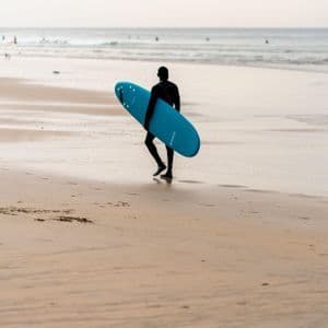 Una persona in muta nera cammina lungo una spiaggia sabbiosa, trasportando una tavola da surf blu verso l'oceano.
