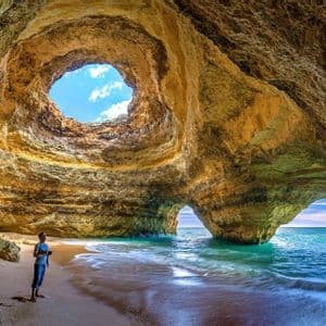 Una persona si trova su una spiaggia sabbiosa all'interno di una grande grotta marina con un oculo nel soffitto e un arco che si apre verso il mare.