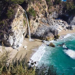 Una cascata scende da una scogliera alberata su una spiaggia sabbiosa che si affaccia su un mare turchese.