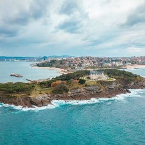 Veduta aerea di un palazzo su una penisola boscosa che si protende nell'oceano, con una città costiera sullo sfondo sotto un cielo nuvoloso.