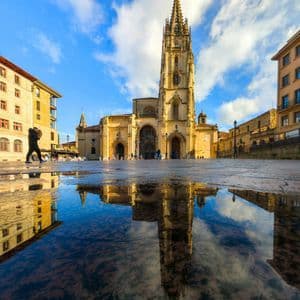 Una vista dal basso di una cattedrale gotica con un'alta guglia riflessa in una grande pozzanghera su una piazza di pietra.