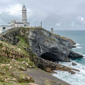 Un faro si erge su una scogliera rocciosa stratificata a picco sul mare, con un viaggio di gruppo WeRoad visibile su un sentiero lungo il fianco della collina.