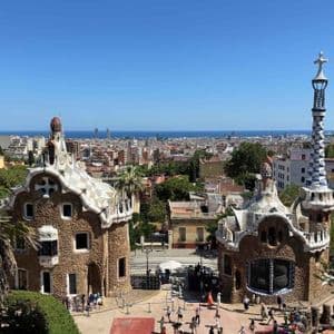 Due stravaganti edifici in pietra con tetti a mosaico si affacciano su un paesaggio urbano che si estende fino al mare sotto un cielo azzurro e limpido.