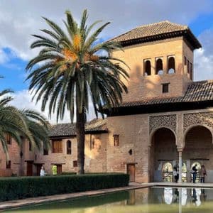 A brick building with arches and a tower next to a reflecting pool and palm trees in a courtyard.